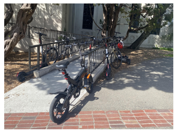 Picture of a wheelchair ramp on the south east corner of VLSB. Several bikes and electric scooters are secured to the handrails, with one blocking the sidewalk access to the ramp.